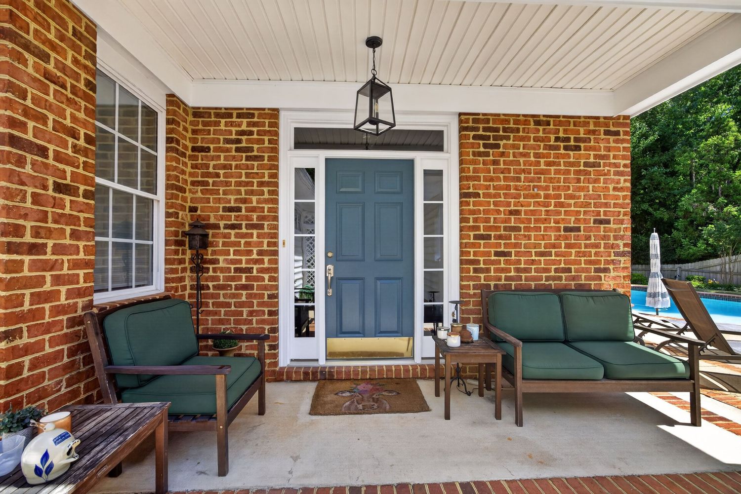 Brick home covered porch with teal door, green cushioned seating, pendant light, and pool glimpse.