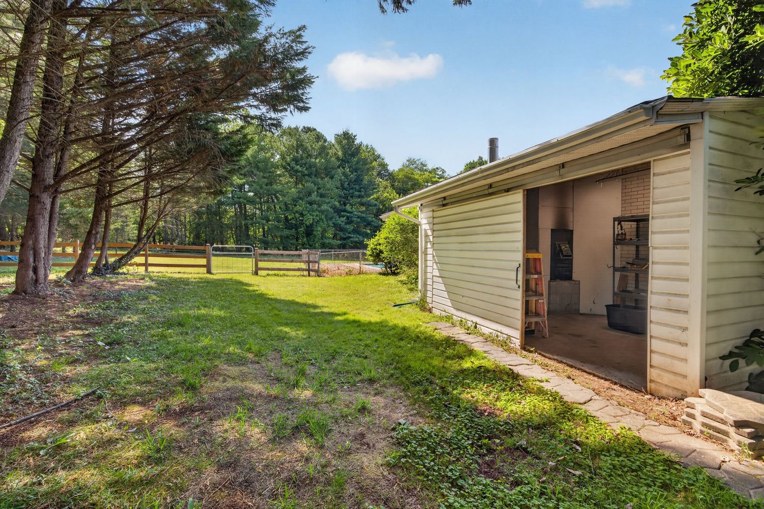 A charming white outbuilding with open doors sits beside lush green lawn and tall trees with a wooden fence in the background.