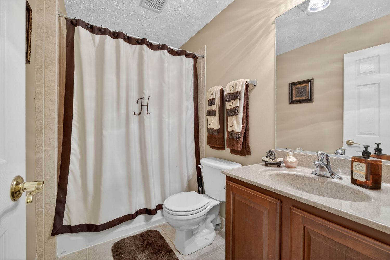 A clean bathroom featuring a monogrammed shower curtain, wooden vanity with mirror, and neutral beige walls.