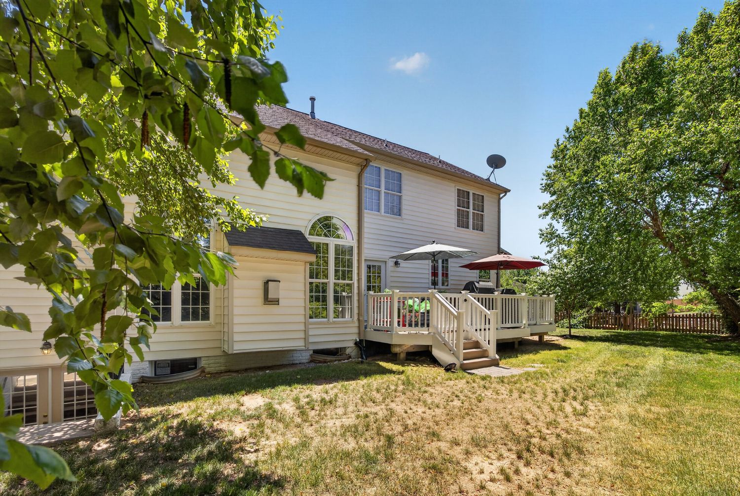 Two-story beige home with large wooden deck, patio umbrella, and lush green yard surrounded by mature trees on a sunny day.