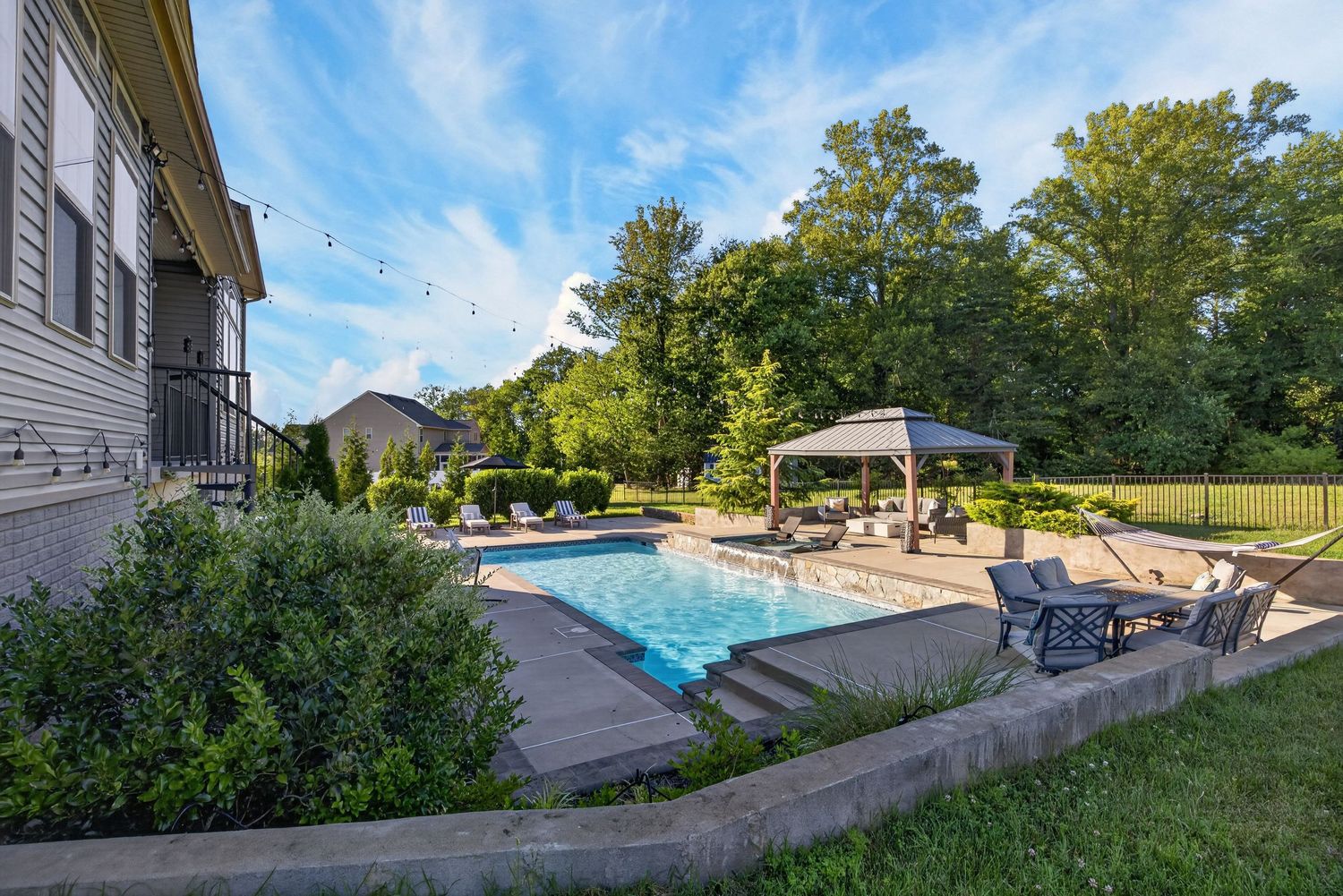 Backyard oasis featuring a rectangular pool, gazebo, patio seating, and lush green lawn surrounded by mature trees.