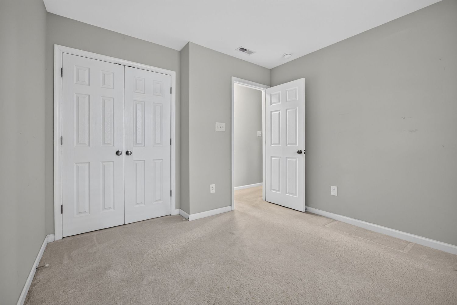 Bright bedroom with light gray walls, beige carpet, white double closet doors, and an open door leading to a hallway.