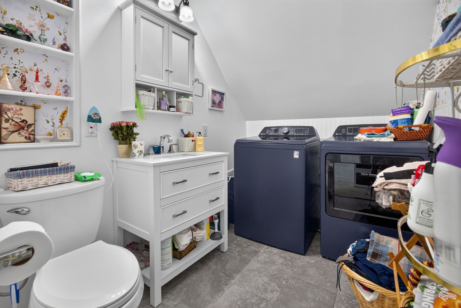 Compact bathroom with white vanity, open shelving, toilet, and navy blue washer and dryer set under sloped ceiling.