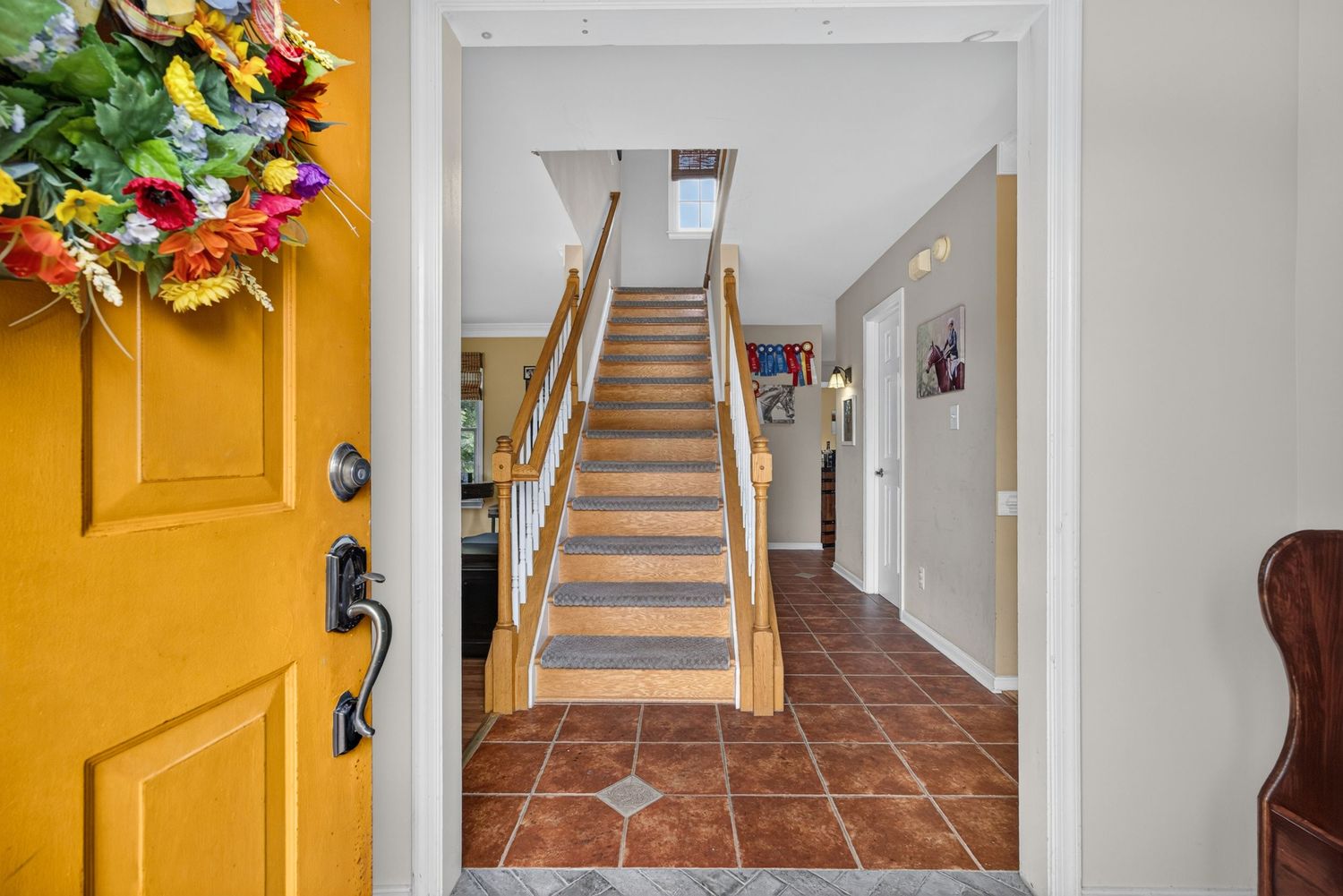 Bright entryway with terracotta tile floors, wooden staircase, skylight, and a colorful floral wreath on a yellow front door.