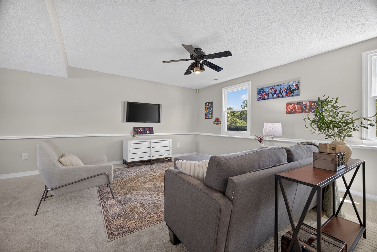 Bright living room with gray sofa, ceiling fan, carpet flooring, white dresser, wall-mounted TV, and large window.
