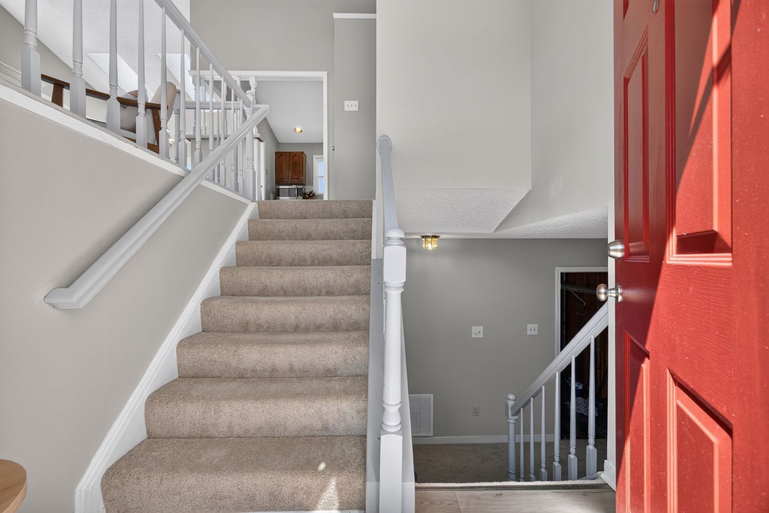 Carpeted staircase with white railings and balusters leads to upper level, featuring a bright red front door and gray walls.