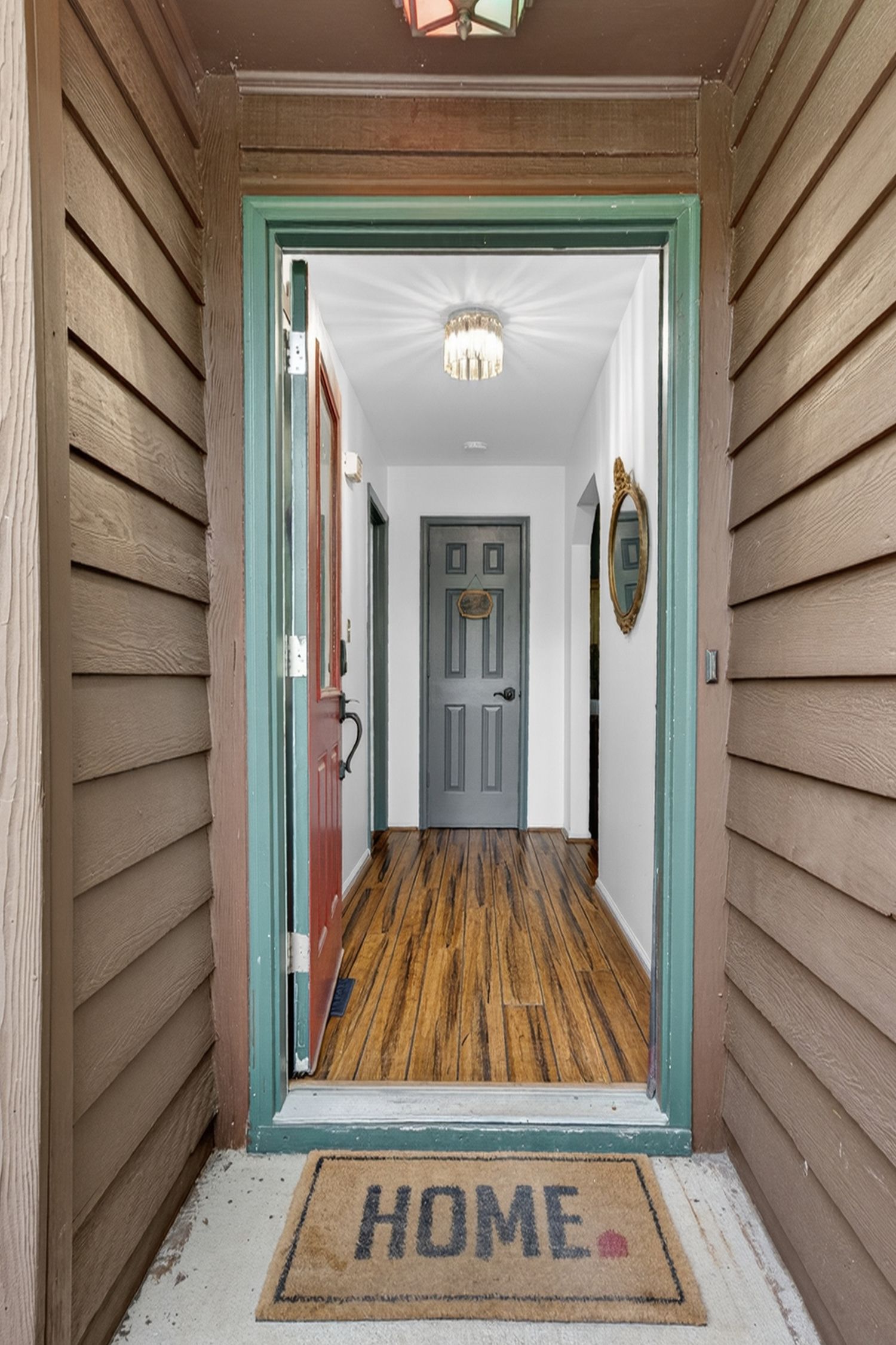 Welcoming home entryway with teal door frame, HOME doormat, hardwood floors, and a hallway leading to a gray interior door.
