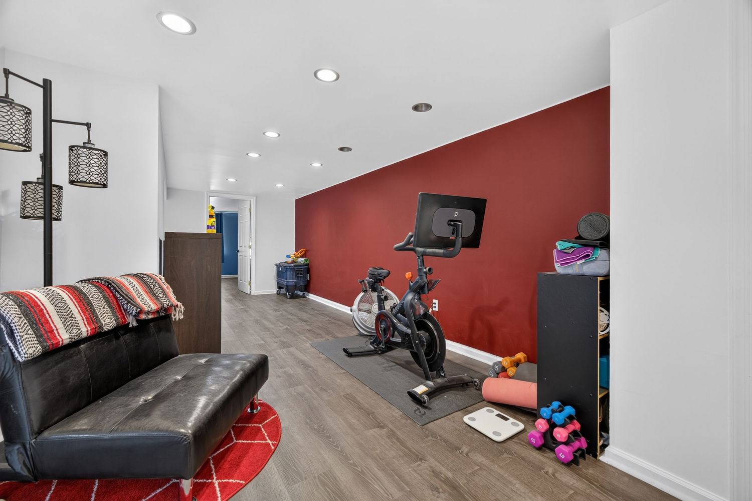 Home gym with red accent wall, Peloton bike, dumbbells, hardwood floors, and a black leather couch.