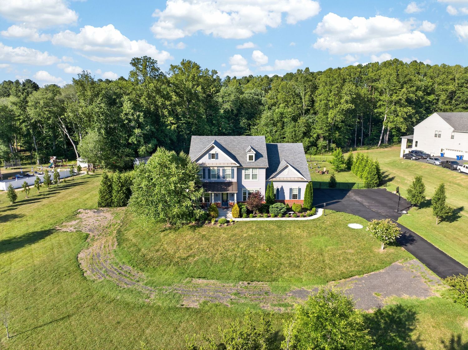 Aerial view of a large two-story home with a circular driveway, lush green lawn, and wooded backdrop on a sunny day.