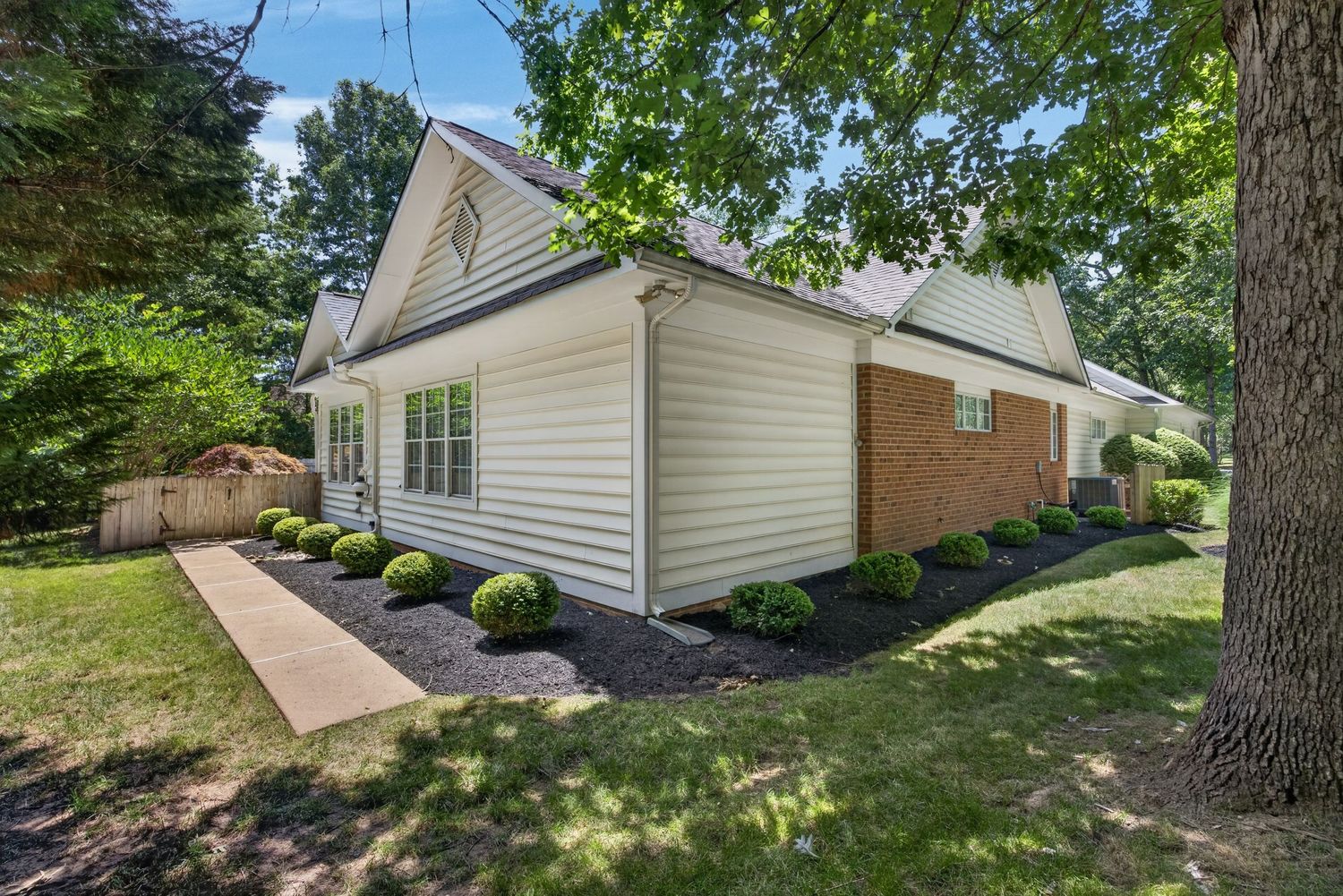 White vinyl siding home with brick accent, lush green landscaping, mature trees, and concrete walkway on a sunny day.