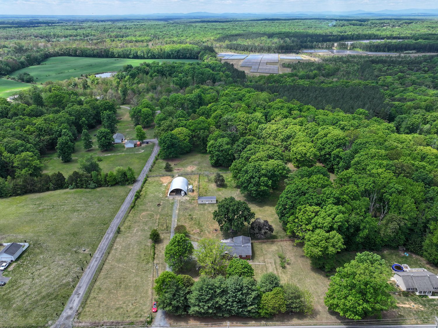 Aerial view of a rural property with a white barn, surrounded by lush green trees, open fields, and a winding road.