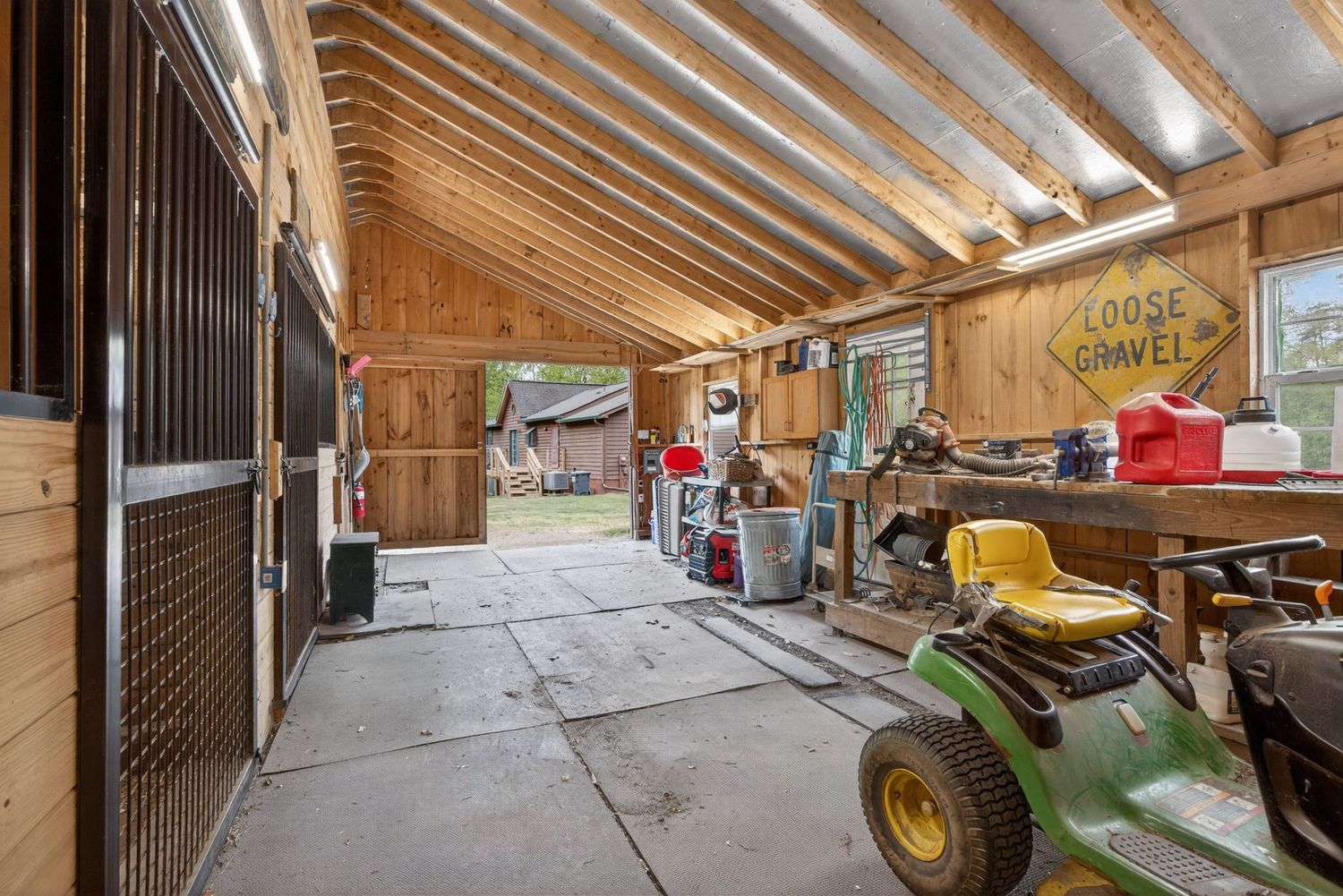 Rustic barn interior with wooden stalls, exposed beam ceiling, green riding mower, and tools stored on a concrete floor.