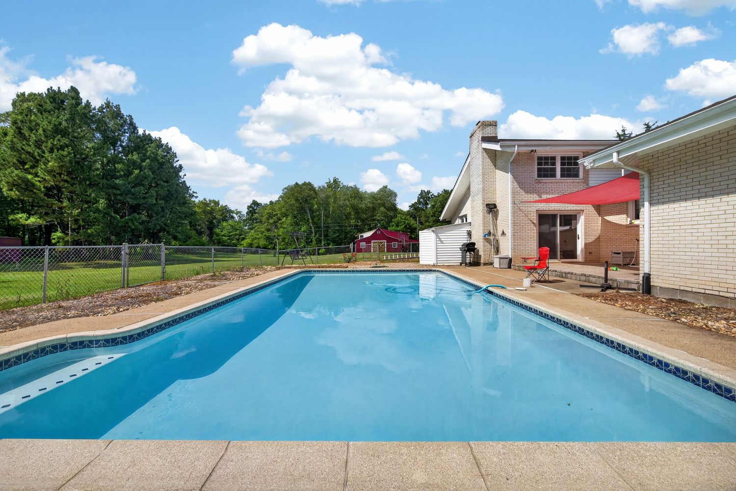 Large rectangular backyard swimming pool with blue water beside a brick home with red umbrella and lush green trees.