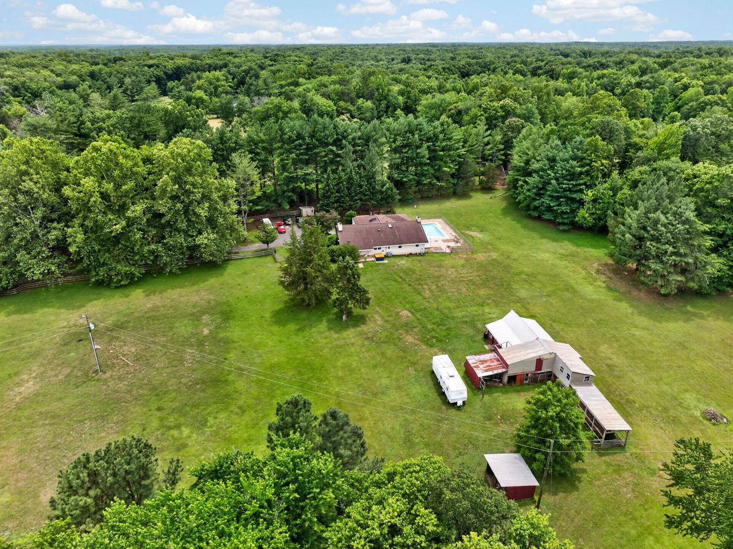 Aerial view of a rural property with a red-roofed home, outbuildings, and open green lawn surrounded by dense woodland.