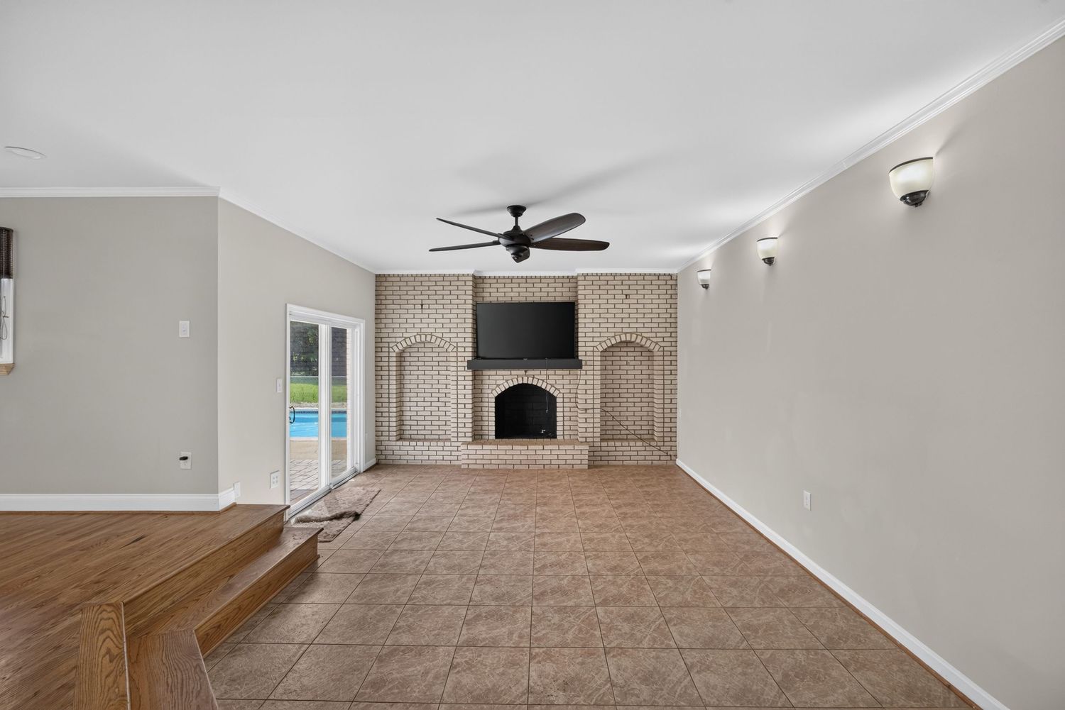 Spacious living room with brick fireplace, wall-mounted TV, ceiling fan, tile floors, and French doors leading to a pool.