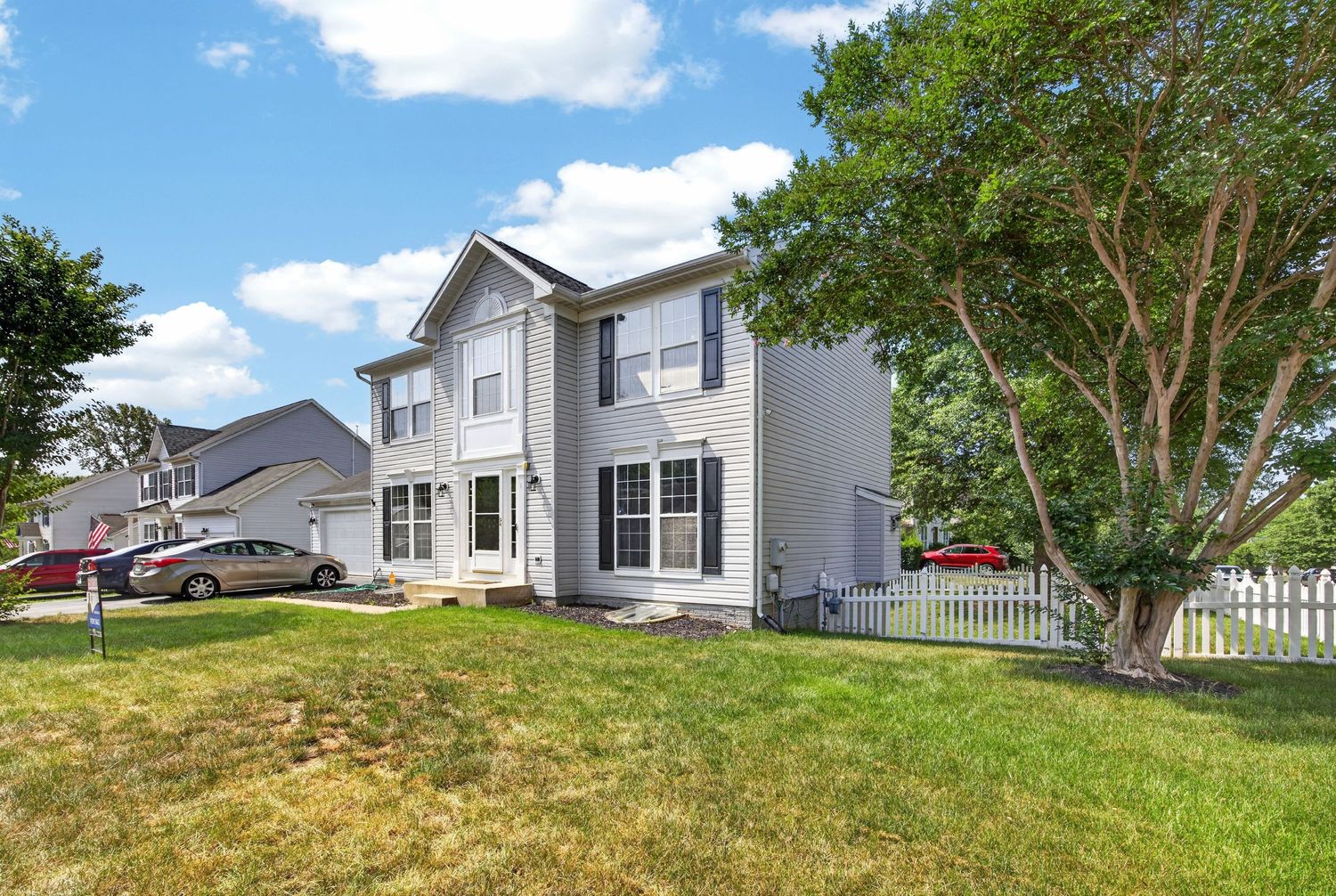 Two-story gray colonial home with white fence, large tree, and well-maintained lawn on a sunny day.