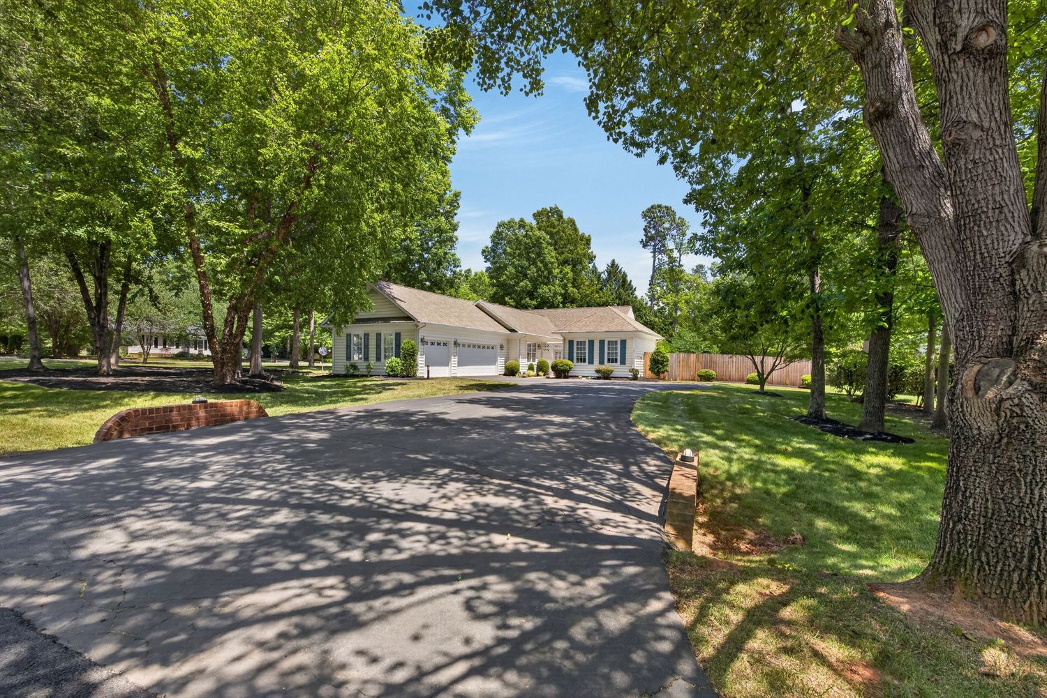 Ranch-style home surrounded by mature trees with a spacious asphalt driveway and lush green lawn on a sunny day.