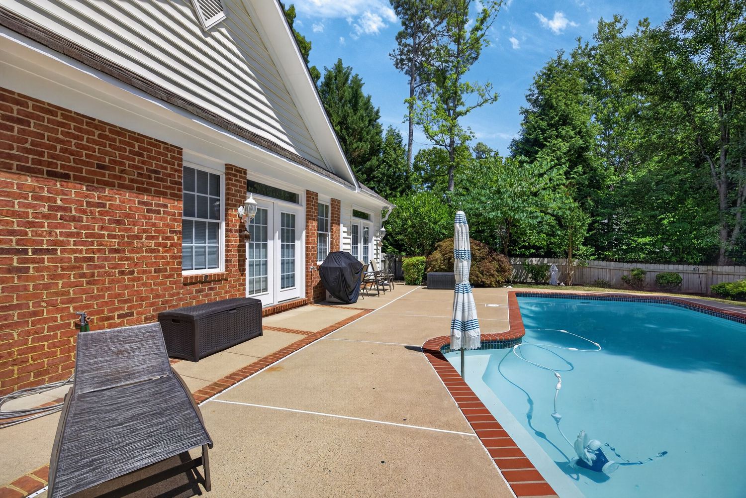 Brick home patio with lounge chairs, BBQ grill, and sparkling blue swimming pool surrounded by lush green trees.