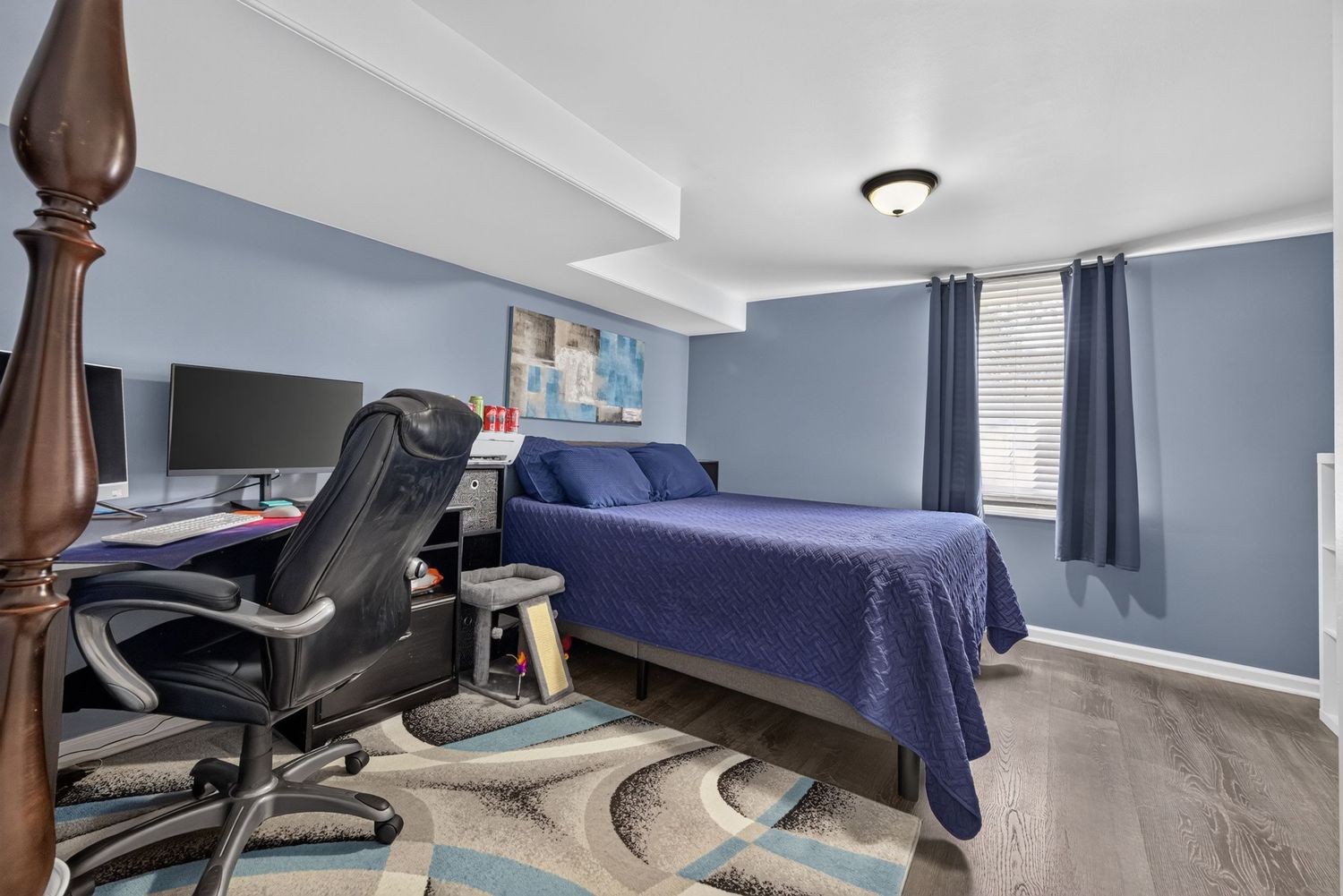 Bedroom with blue walls, hardwood floors, navy bedding, desk setup, and teal curtains on a bright window.