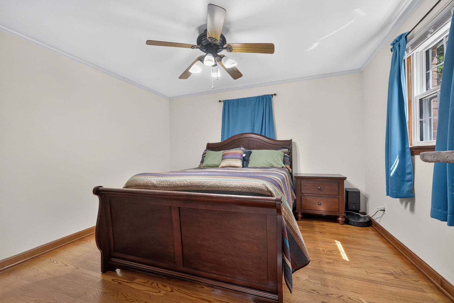 Cozy bedroom with dark wood sleigh bed, matching dresser, hardwood floors, ceiling fan, and blue curtains on windows.
