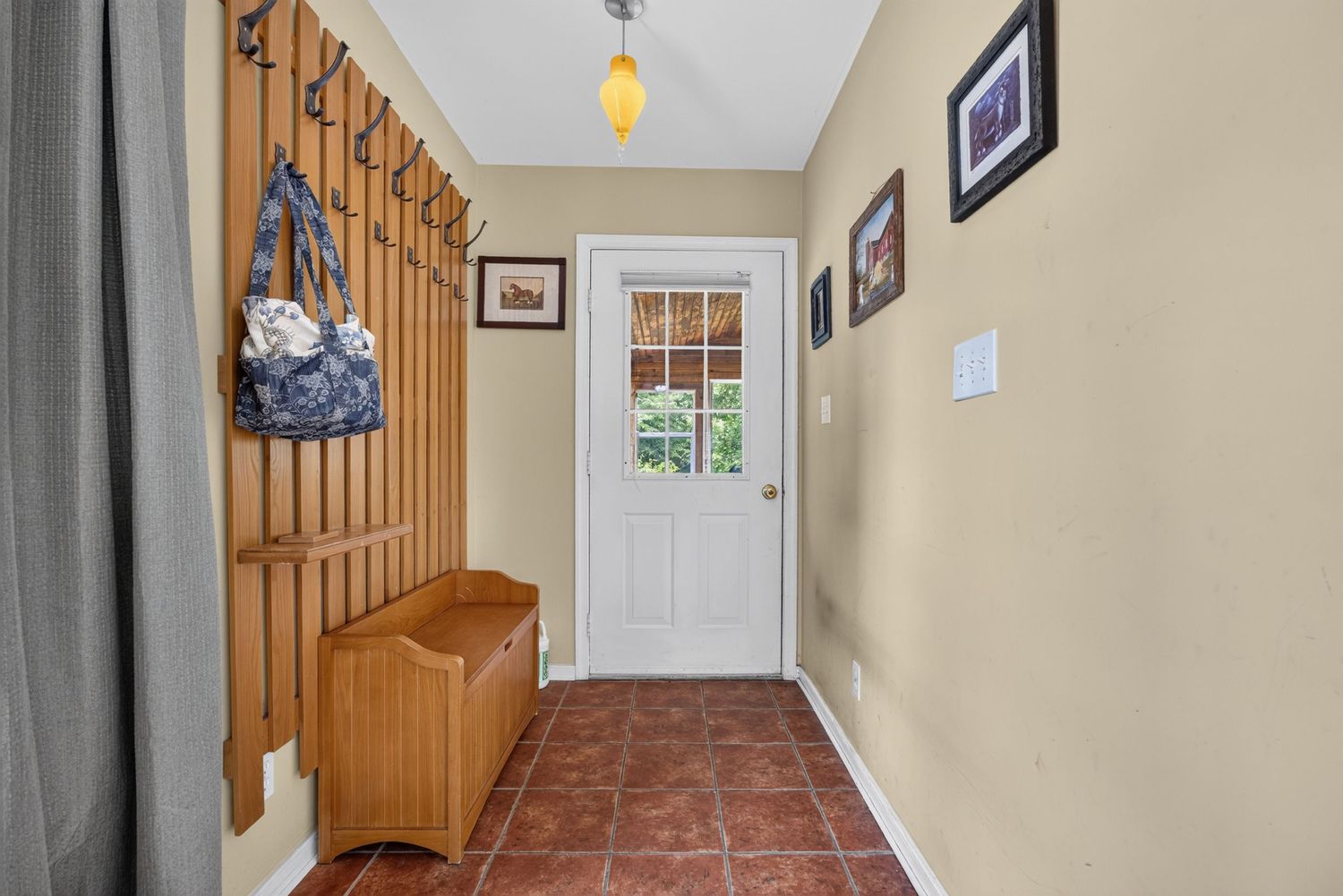 Mudroom entryway with terracotta tile floor, wooden coat rack with hooks, storage bench, and white door with window.