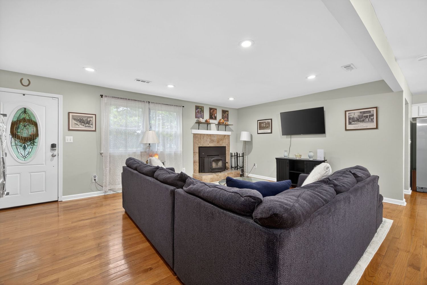 Bright living room with gray sectional sofa, hardwood floors, fireplace, wall-mounted TV, and recessed lighting.