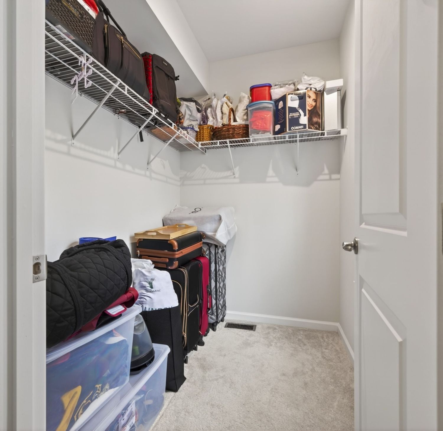 Walk-in closet with wire shelving, carpet flooring, storage bins, and luggage neatly organized along the walls.