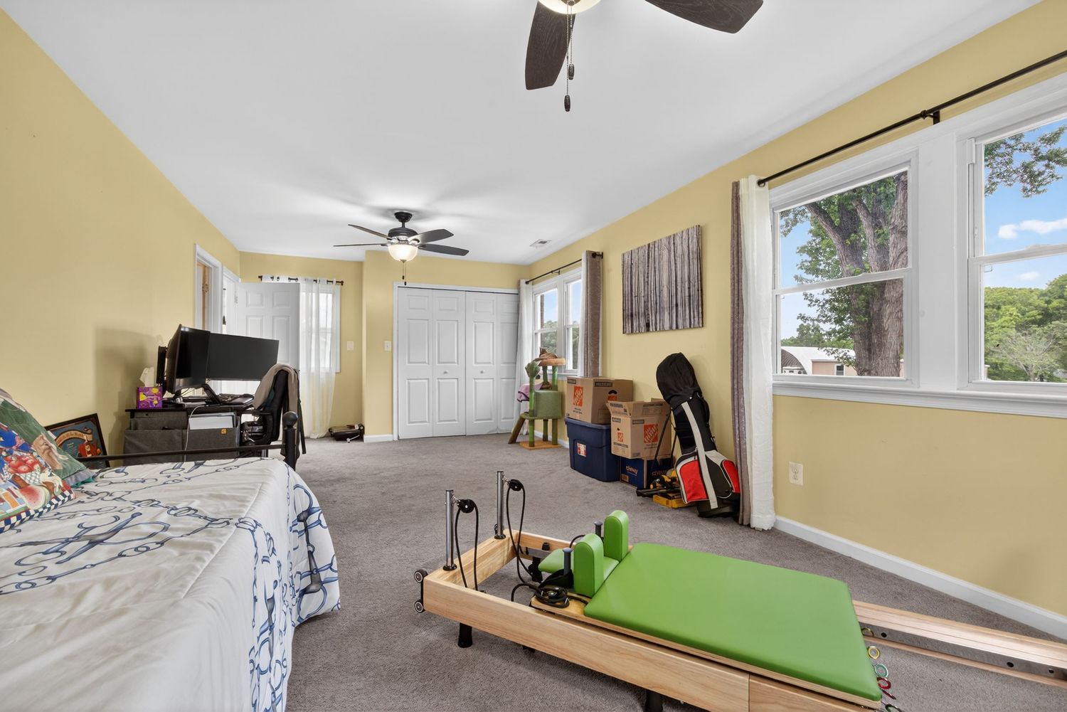Spacious bedroom with gray carpet, ceiling fan, large windows, a bed, and exercise equipment on the floor.