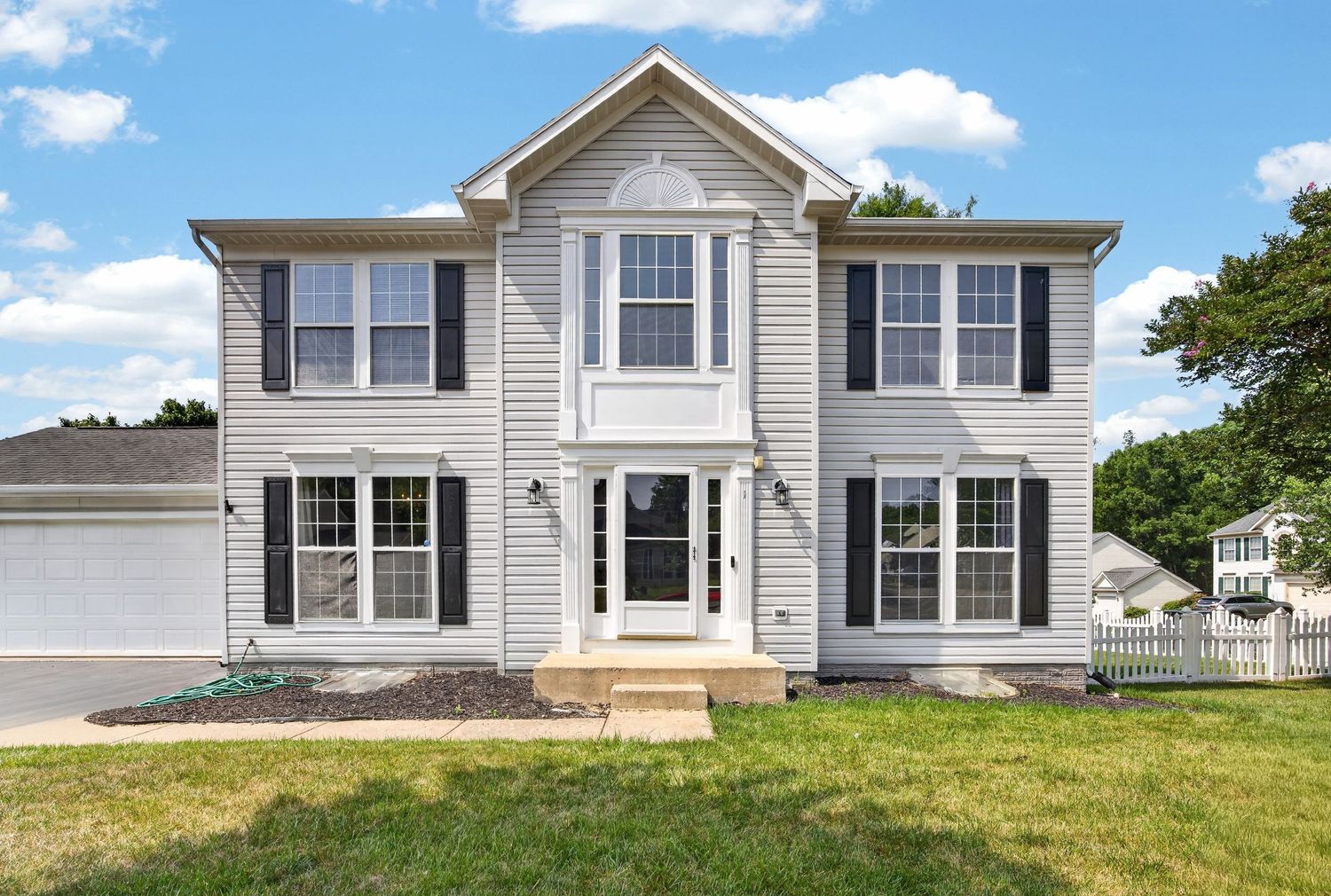 Two-story colonial home with gray siding, black shutters, white entry door, attached garage, and lush green lawn under blue skies.
