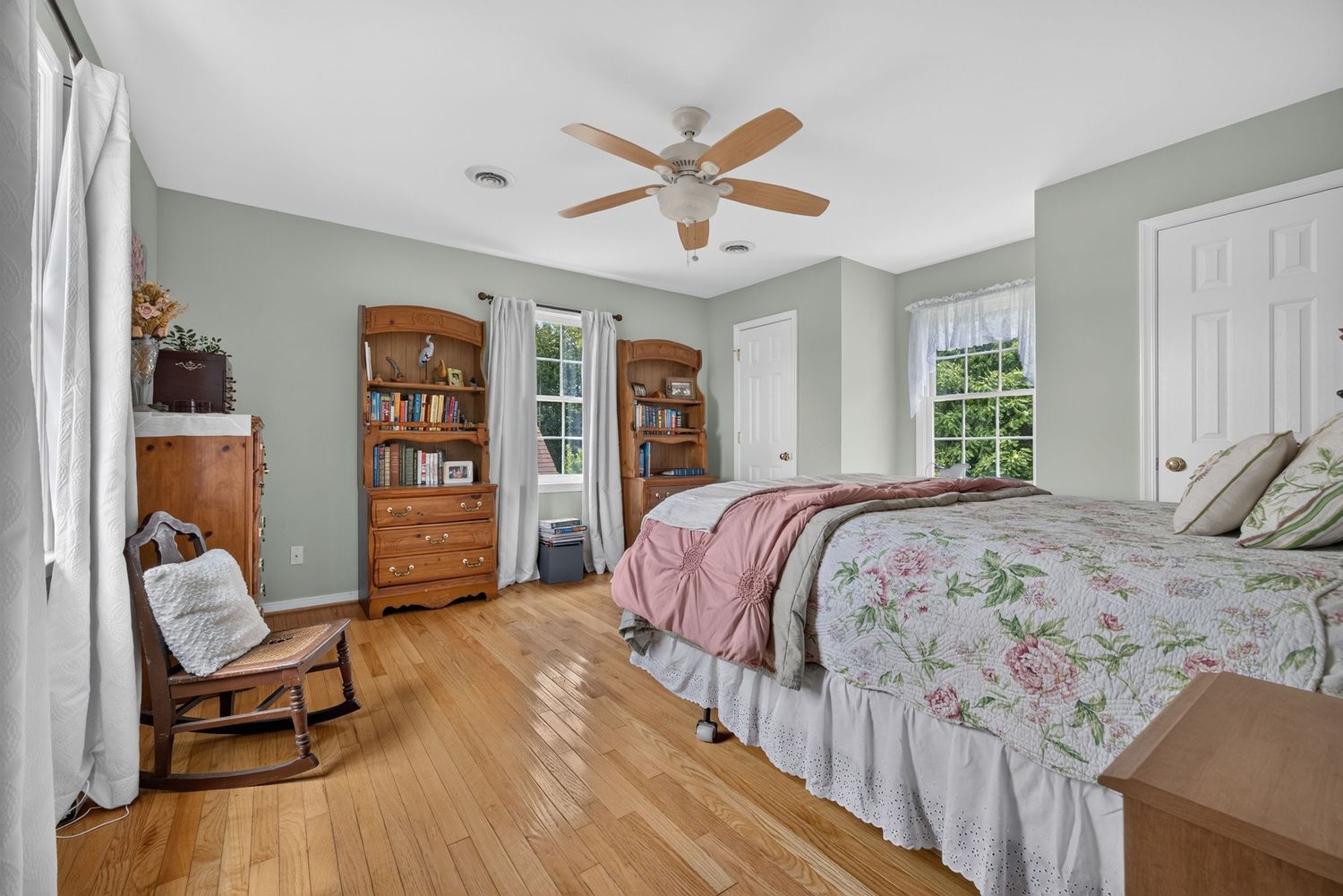 Spacious bedroom with hardwood floors, sage green walls, floral bedding, rocking chair, wooden bookcase, and ceiling fan.