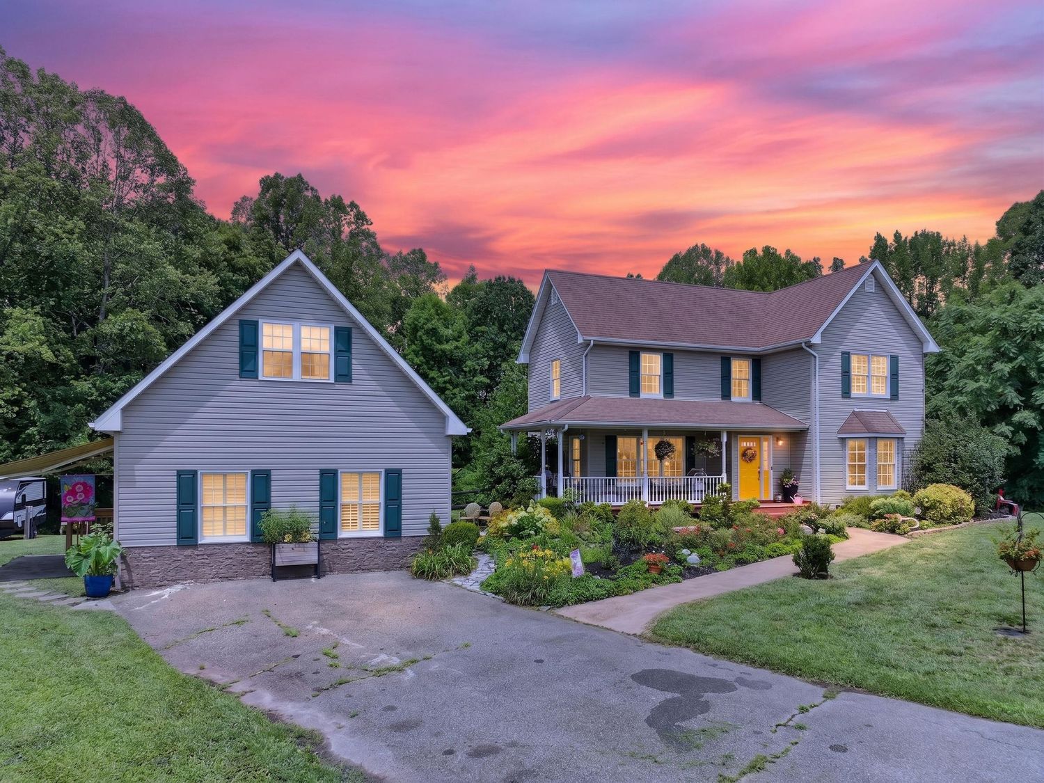 Two-story gray colonial home with green shutters and covered porch glowing warmly beneath a vibrant pink and purple sunset sky.