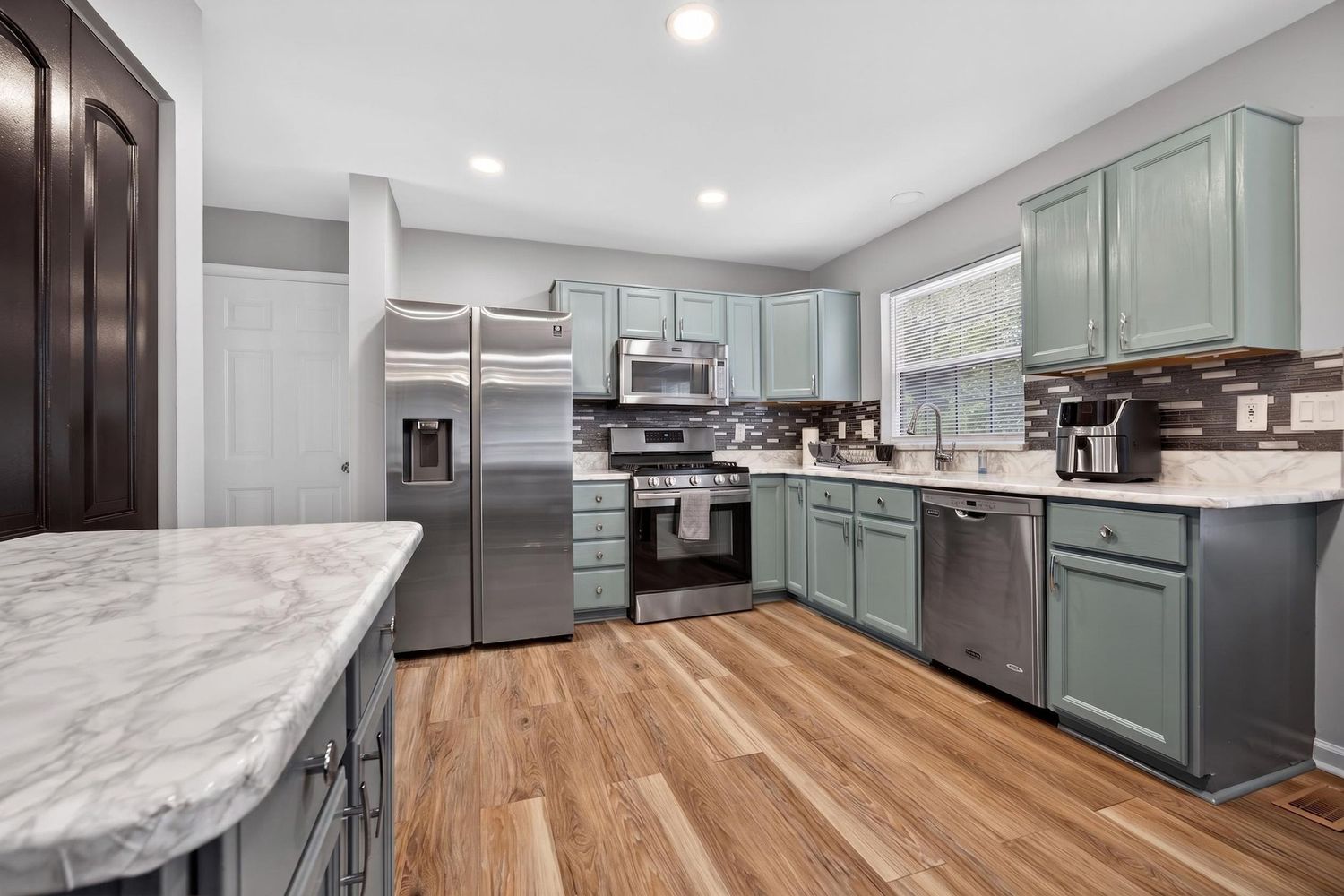 Modern kitchen with sage green cabinets, stainless steel appliances, marble countertops, and hardwood floors.
