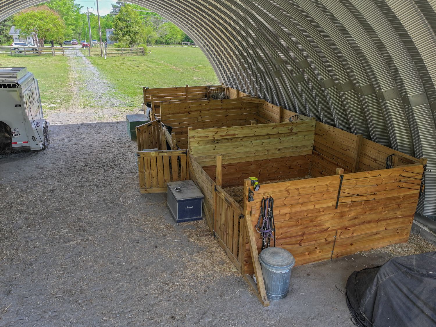 Wooden compost bins inside a metal quonset hut shelter on a gravel surface with green grass visible outside.