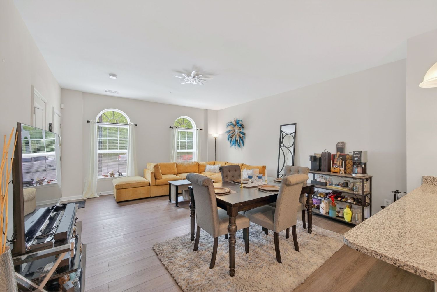 Open-concept living and dining room with hardwood floors, yellow sofa, arched windows, and granite countertop accent.