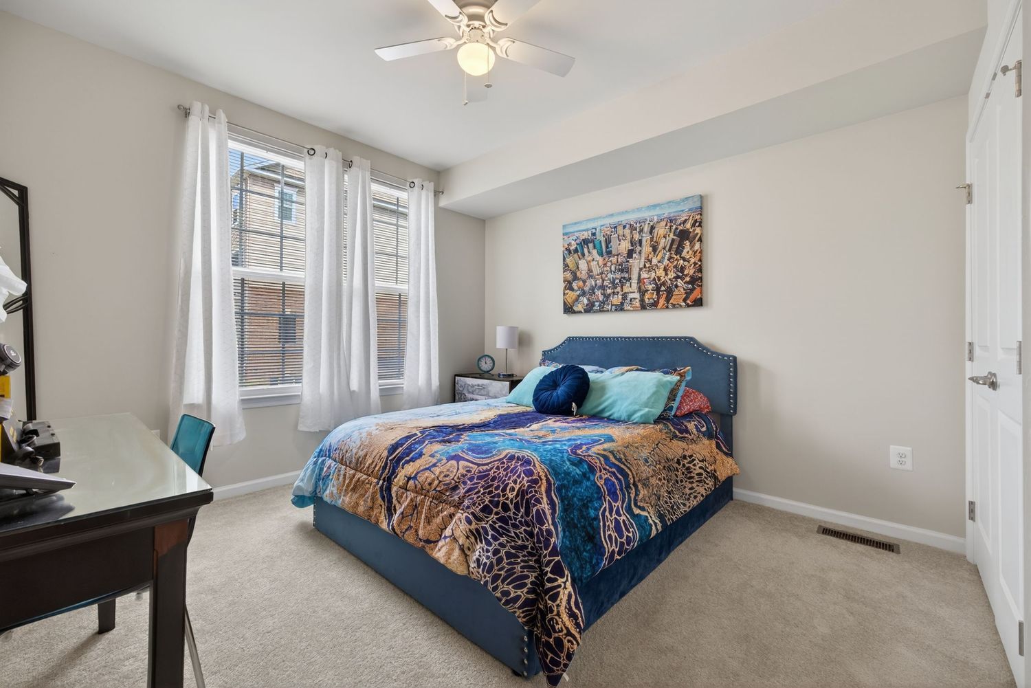 Cozy bedroom with blue upholstered bed, colorful bedding, carpet flooring, ceiling fan, and city skyline artwork above the bed.
