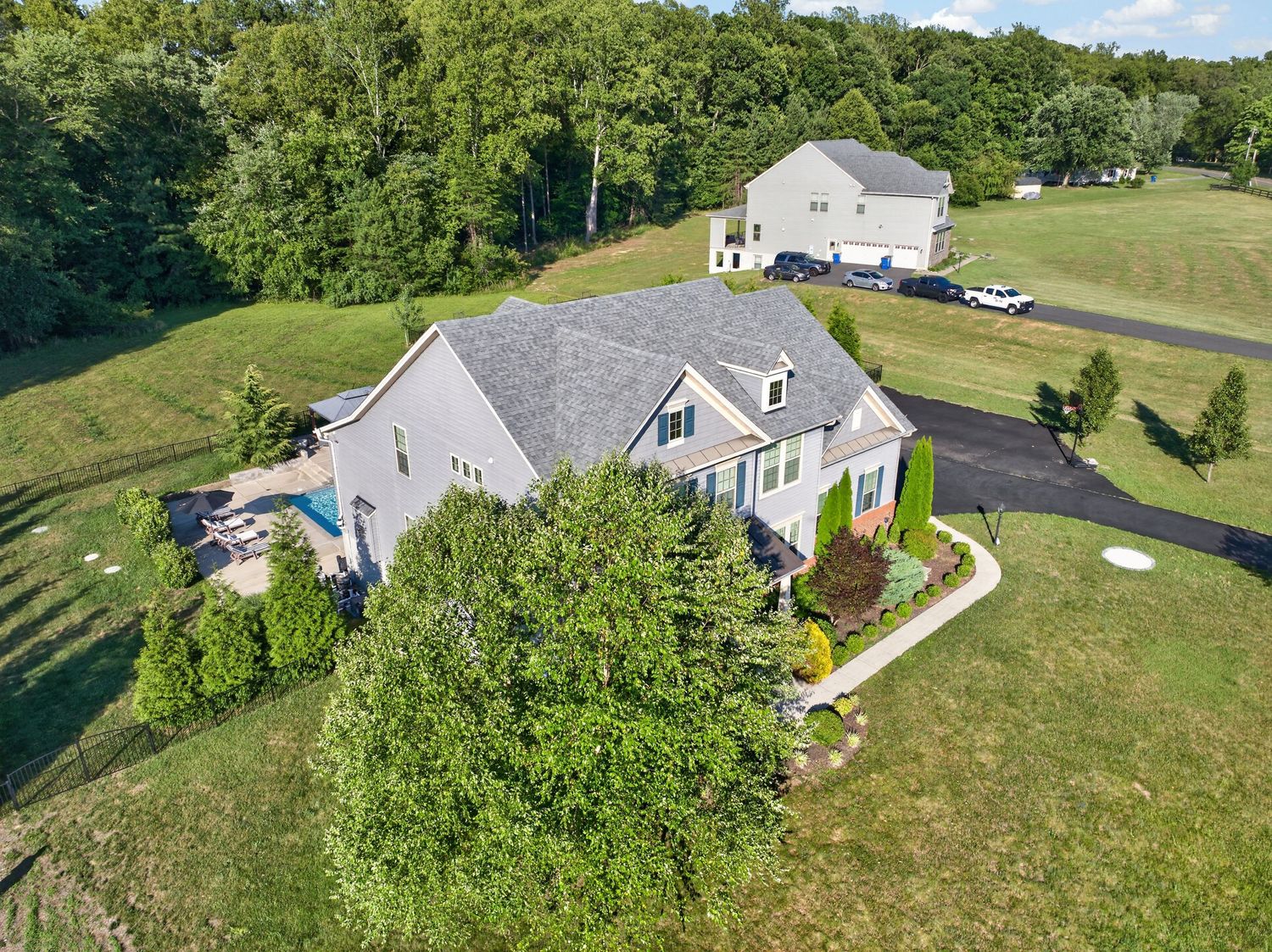 Aerial view of a large suburban home with lush green lawn, mature trees, swimming pool, and curved driveway in a rural setting.