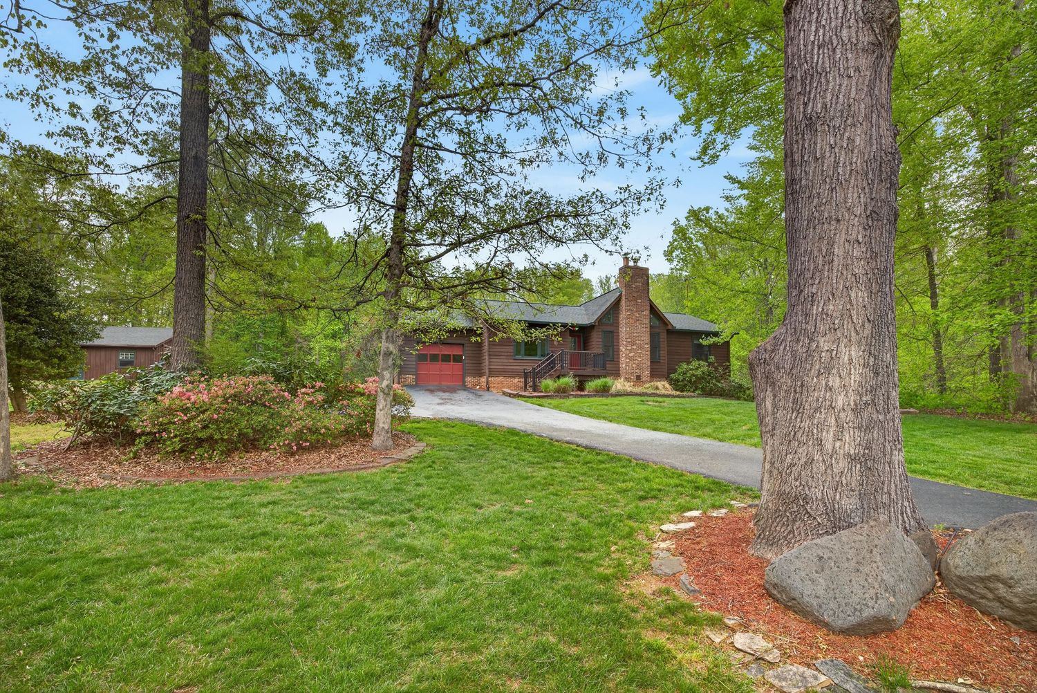 Charming brick ranch home surrounded by tall trees and lush green lawn with a curved driveway in a wooded setting.