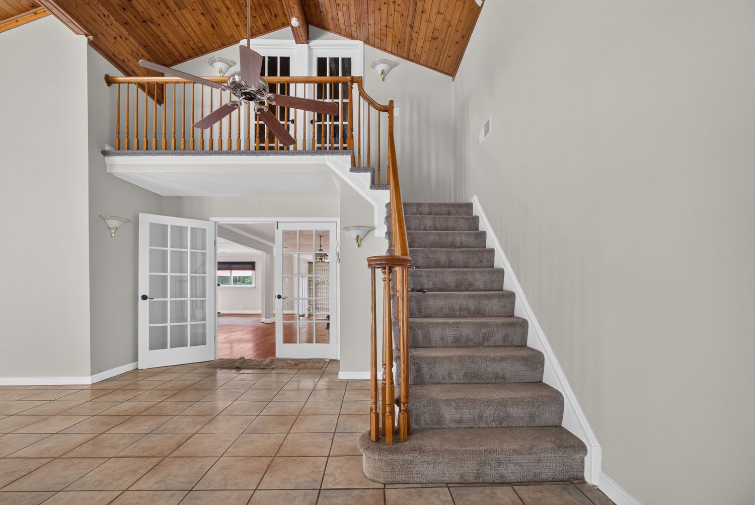 Spacious foyer with tile floors, carpeted staircase, wood railings, vaulted ceiling, and French doors leading to a bright living area.