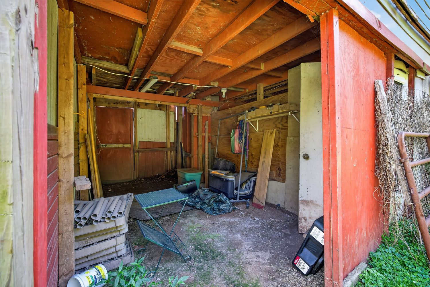 Rustic red wooden shed interior with exposed beam ceiling, scattered tools, and overgrown vegetation at the entrance.