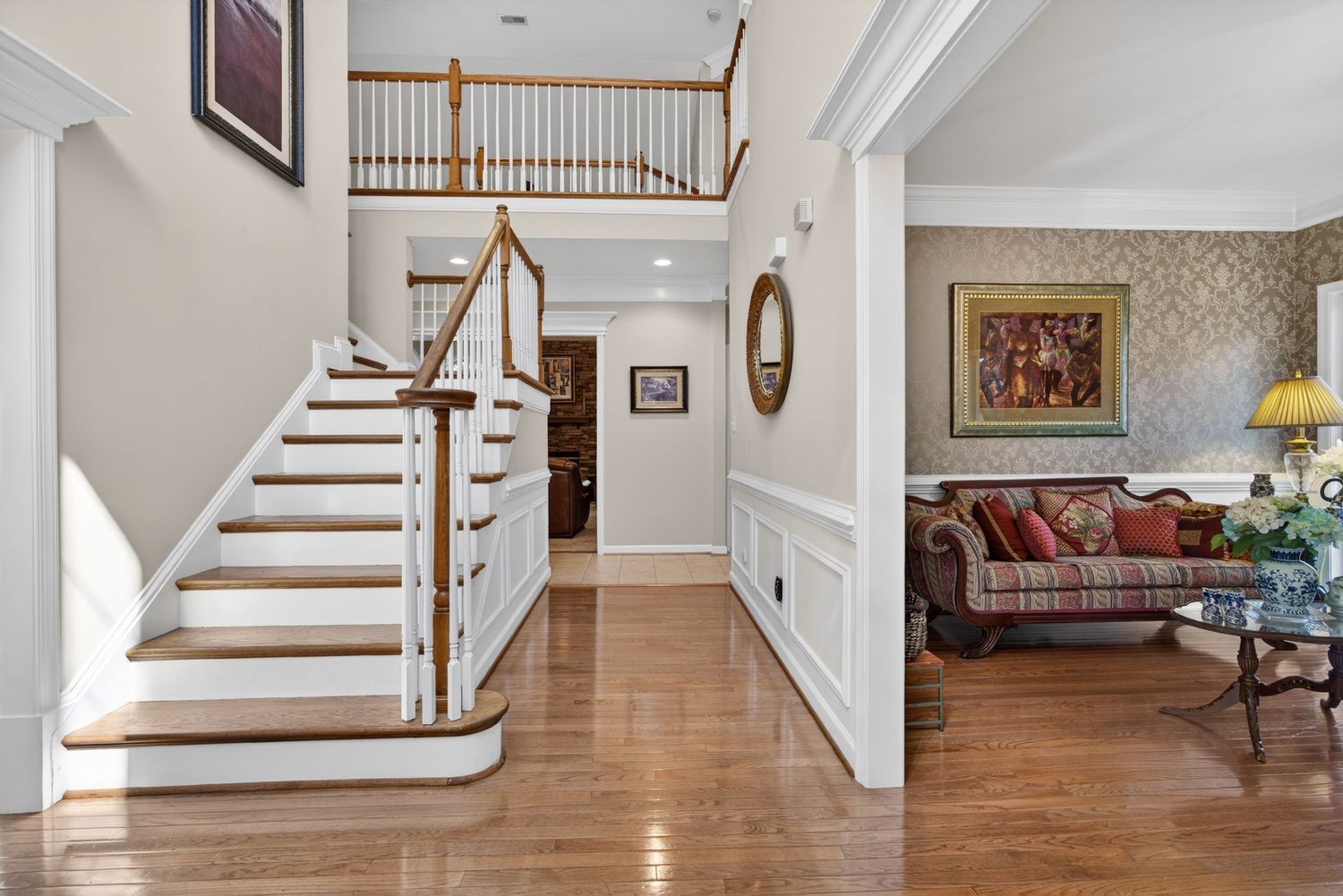 Elegant two-story foyer with hardwood floors, white staircase, and view into a traditional living room with floral wallpaper.