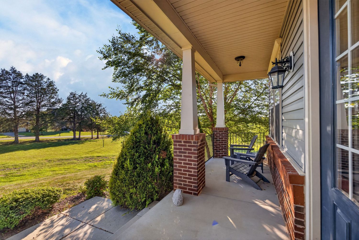 Covered front porch with brick columns overlooking a lush green lawn and mature trees on a sunny day.
