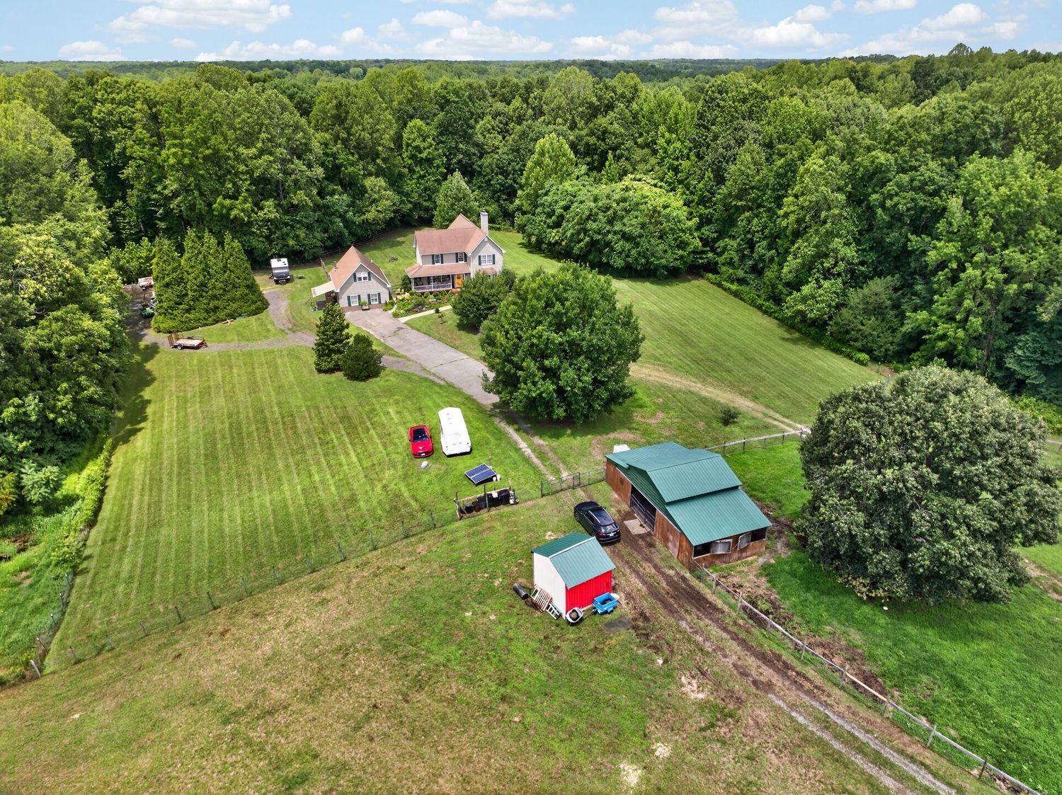 Aerial view of a rural property featuring a pink farmhouse surrounded by lush green trees, open lawn, and red farm buildings.