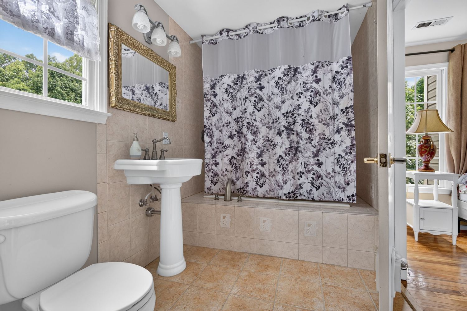 Bright bathroom with pedestal sink, toilet, tiled walls, and a bathtub with floral shower curtain near a window.