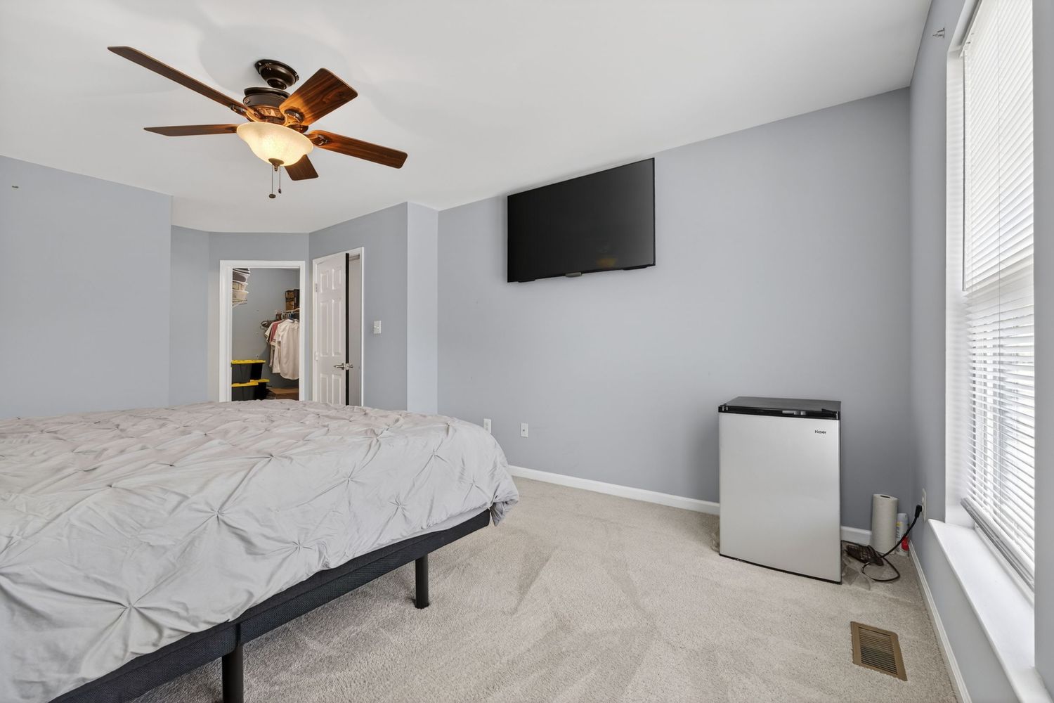 Spacious bedroom with gray walls, ceiling fan, wall-mounted TV, carpet flooring, large bed, and natural light from window.