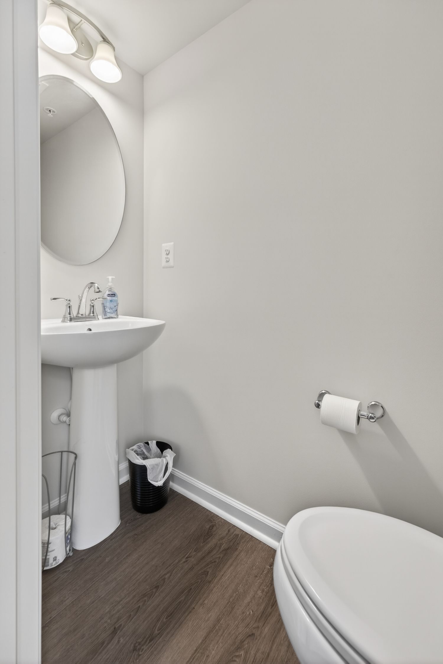 Modern powder room with pedestal sink, round mirror, wood-look floors, white walls, and toilet with chrome paper holder.