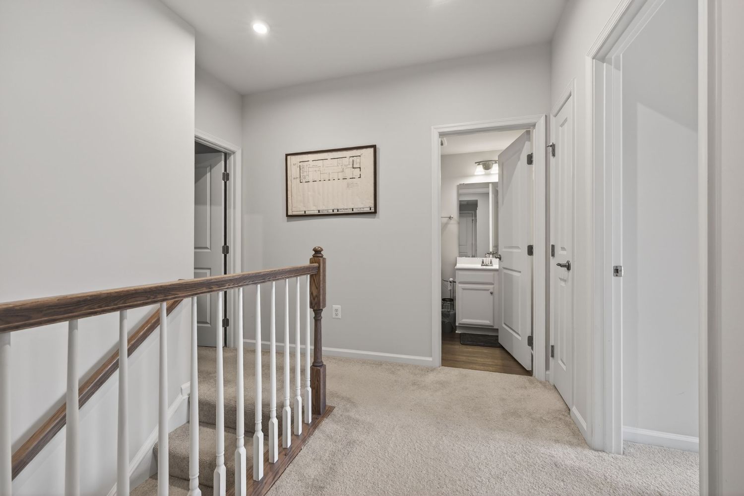 Bright upstairs hallway with beige carpet, white railings, and a view into a bathroom at the end of the corridor.