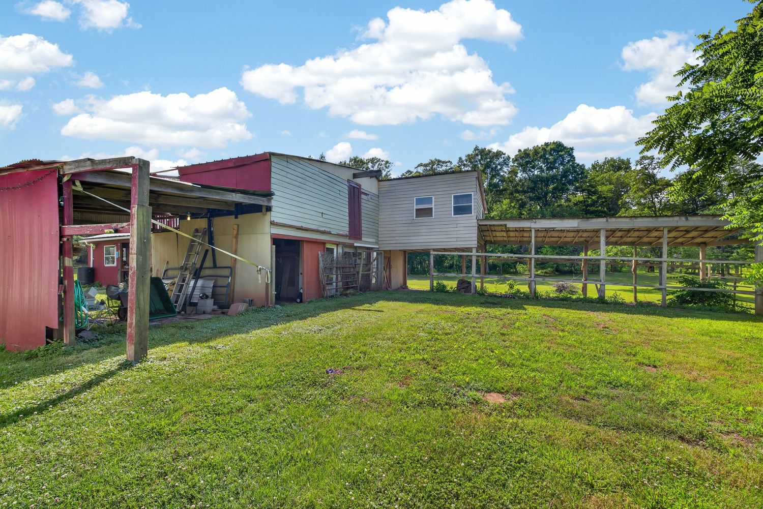 Rural property featuring a two-story house with green lawn, red outbuilding, and long covered animal pen under a blue sky.