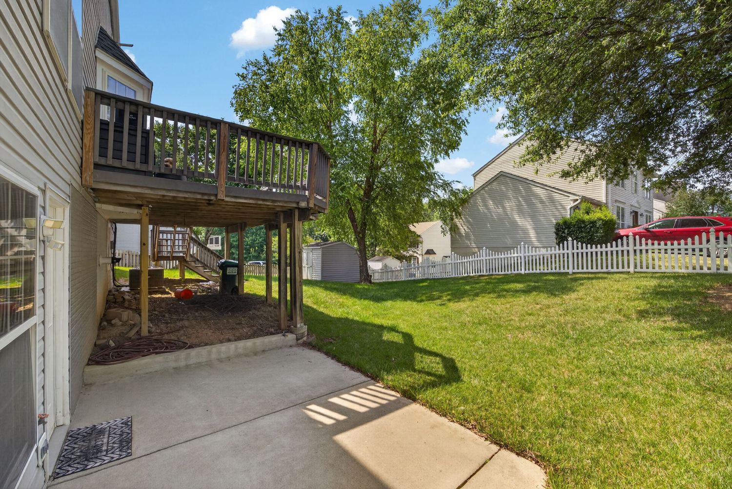 Spacious backyard with lush green lawn, elevated wooden deck, patio, mature trees, and white privacy fence.
