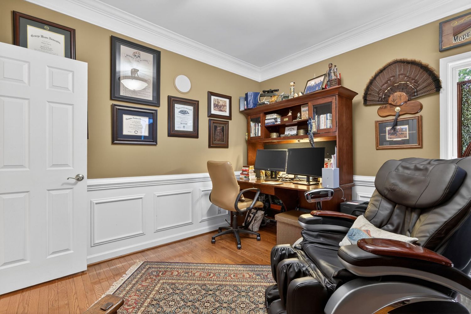 Home office with wainscoting, hardwood floors, mahogany desk, massage chair, and framed wall art.