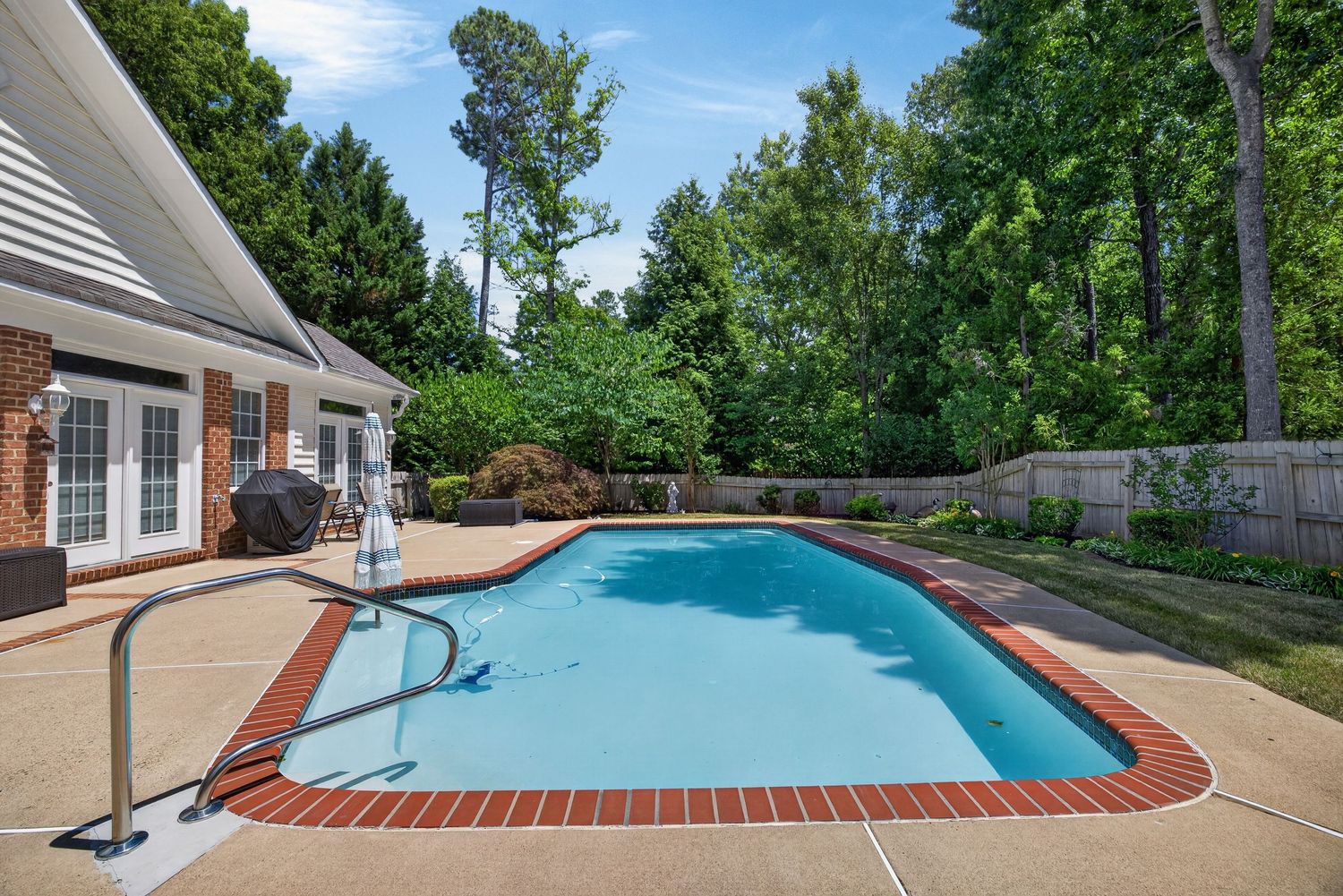 Backyard swimming pool with brick border, patio, and lush green trees in a fenced suburban yard.